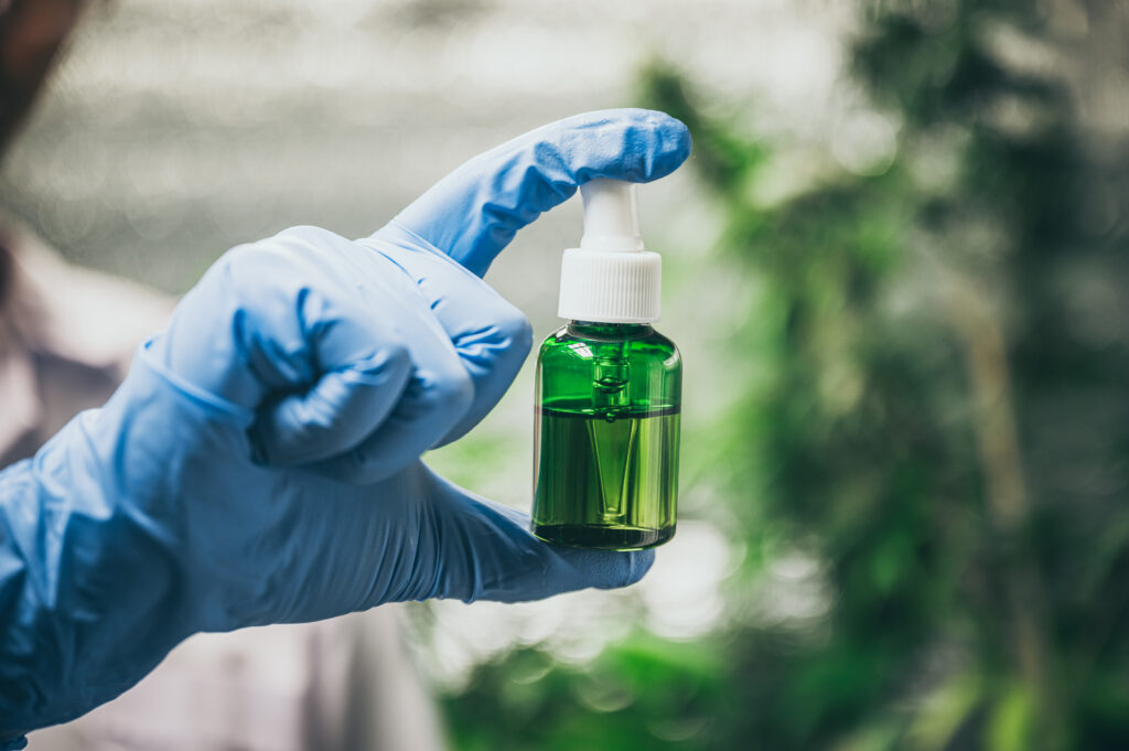 Mão com luva azul segurando frasco conta-gotas de vidro verde contendo óleo de canabidiol (CBD), com fundo desfocado de plantas, representando pesquisa científica sobre terapias naturais.