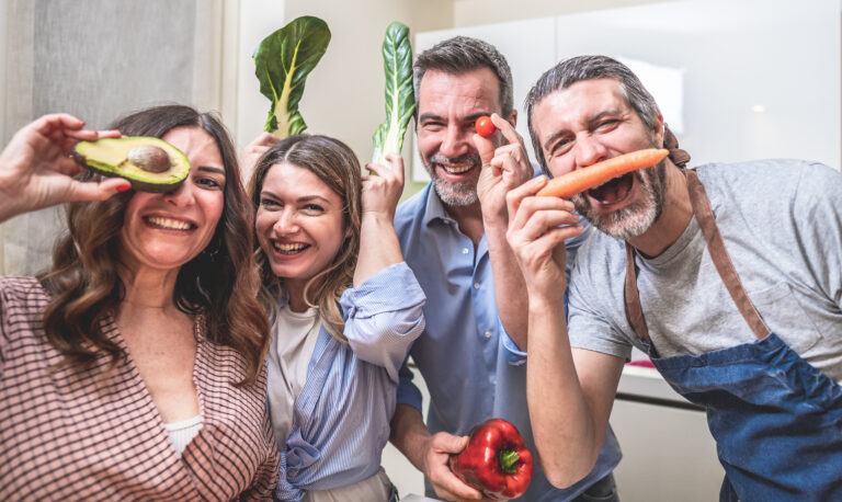 Grupo de adultos sorrindo na cozinha segurando vegetais frescos como abacate, cenoura, acelga, pimentão e tomate, simbolizando uma alimentação saudável e divertida.