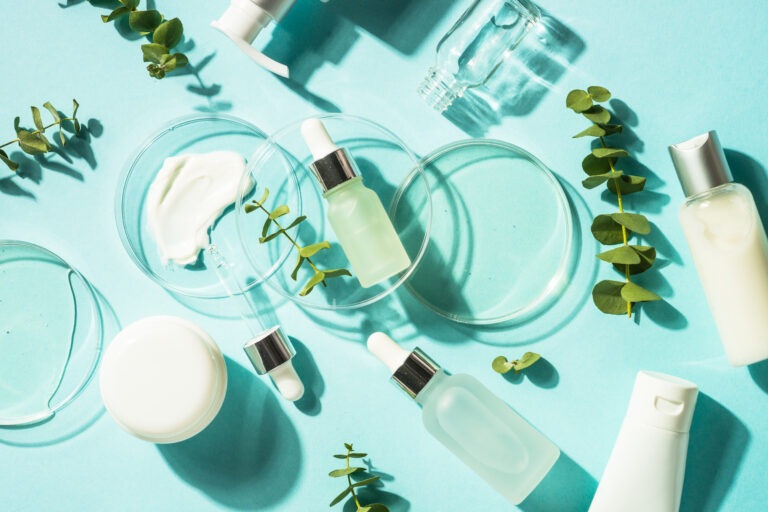 frascos e embalagens de produtos de skincare sobre superfície azul, com placas de Petri, séruns, cremes e folhas verdes, representando ingredientes cosméticos de origem biotecnológica e natural